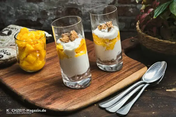 Zwei Gläser mit einem cremigen Mango-Dessert, geschichtet mit zerbröselten Keksen und garniert mit Mango-Stücken. Daneben ein Glas voll frischer Mango-Würfel.