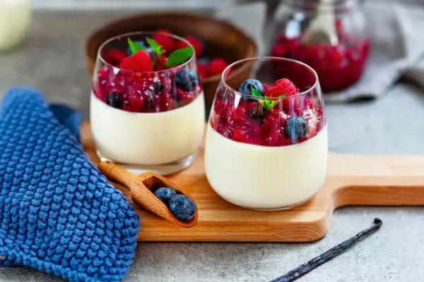 Zwei Gläser cremiger Panna cotta mit Beerenkompott und frischen Beeren, garniert mit Minze. Ein Löffel mit Blaubeeren liegt daneben.