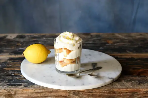 Ein Glas mit geschichtetem Zitronen Tiramisu, garniert mit Zitronenschale. Daneben liegt eine Zitrone.