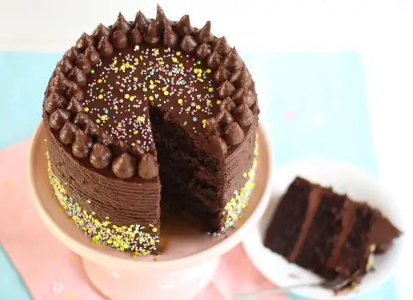 Eine köstliche Schokocreme Torte mit Schokoglasur, Schokotuffs und bunten Streuseln. Ein Stück ist bereits auf einem Teller angerichtet.