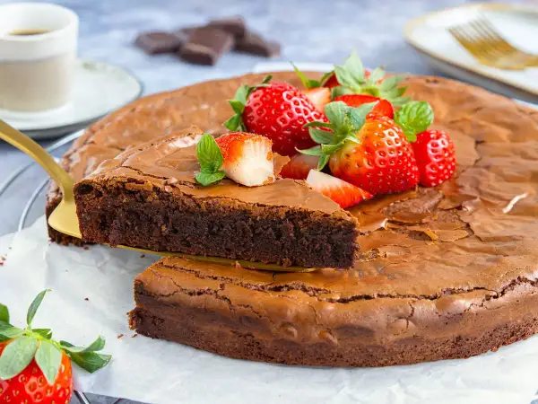 Ein saftiger brownie kuchen, garniert mit frischen Erdbeeren und Minze. Ein Stück wird mit einem goldenen Tortenheber herausgehoben.